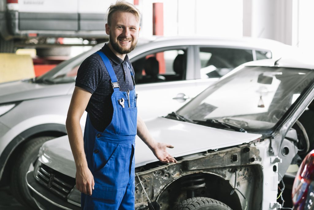 Uśmiechnięty mechanik pracujący przy uszkodzonym samochodzie po kolizji w warsztacie – naprawa powypadkowa.
