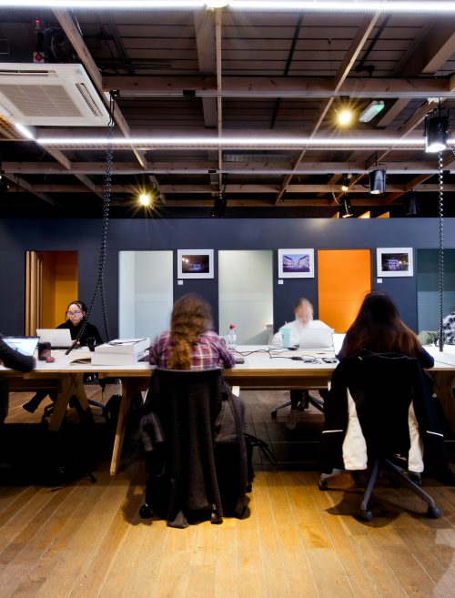 Grupa osób siedzi przy długim stole w nowoczesnym wnętrzu i pracuje przy laptopach