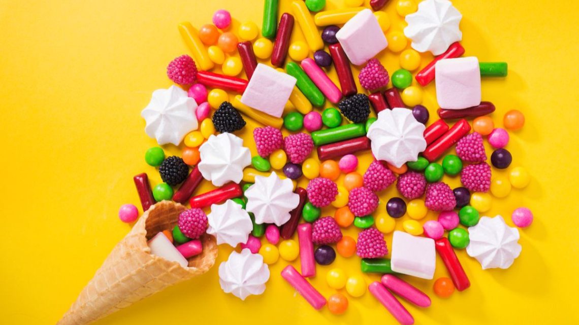 Kolorowe cukierki reklamowe, różowe pianki marshmallow i żelki w kształcie malin wysypane z waflowego rożka na intensywnie żółte tło.