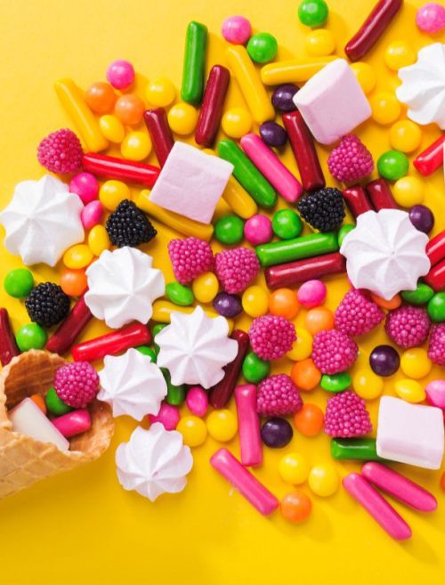 Kolorowe cukierki reklamowe, różowe pianki marshmallow i żelki w kształcie malin wysypane z waflowego rożka na intensywnie żółte tło.