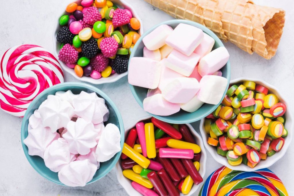 Różnorodne cukierki reklamowe, pianki i lizaki ułożone w miseczkach na jasnym tle; zestaw zawiera kolorowe landrynki, żelki owocowe i różowe bezy.