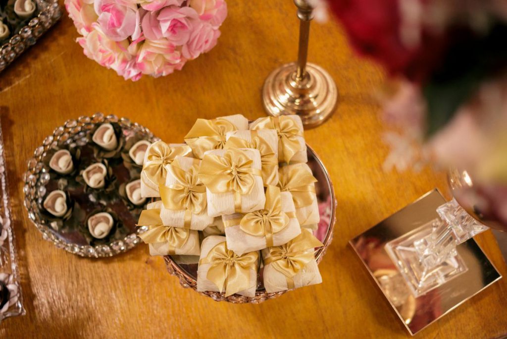 Elegancko zapakowane słodycze na święta w formie małych upominków z kremowego papieru ze złotymi wstążkami, ułożone na ozdobnej paterze obok dekoracji kwiatowych.