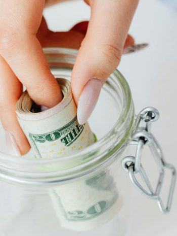 Osoba wkładająca zwinięte banknoty dolarowe do szklanego słoika, co ilustruje domowy program oszczędnościowy i gromadzenie kapitału.