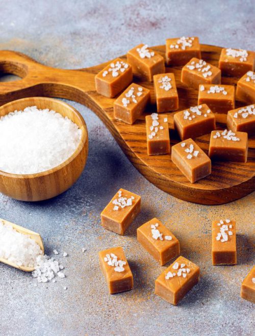 Ręcznie robione krówki z logo posypane kryształkami soli morskiej, ułożone na drewnianej desce obok miseczki z solą; eleganckie i smaczne słodycze personalizowane dla firm.