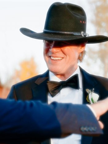 Dwóch elegancko ubranych mężczyzn w kapeluszach rozmawia na zewnątrz podczas uroczystości, w tle widać jesienne drzewa.