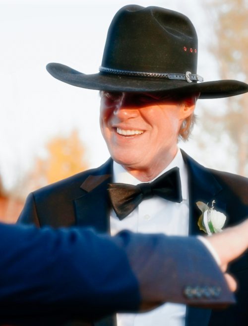 Dwóch elegancko ubranych mężczyzn w kapeluszach rozmawia na zewnątrz podczas uroczystości, w tle widać jesienne drzewa.